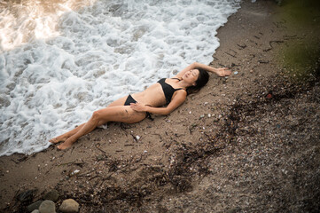 Asian woman in black swimsuit lying on sandy beach at sunset, waves touching her body among stones,...