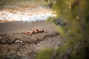 Asian woman in black swimsuit lying on sandy beach at sunset, waves touching her body among stones,...