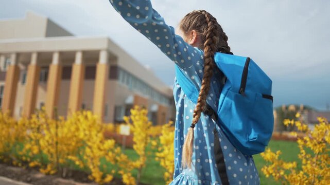 Girl running on campus lawn among yellow shrub and flower near school building carrying blue backpack with long braid swinging in wind showing playful motion in spring urban campus landscape