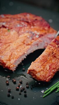 Close-up of juicy grilled pork ribs being sliced with a knife, sprinkled with coarse salt and herbs, served with cherry tomato on black slate.