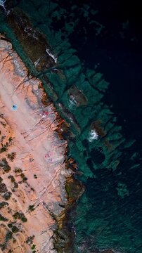 Drone video showing rugged Spanish coastline with turquoise sea, clear shallow waters, and small groups of people relaxing on the rocks.