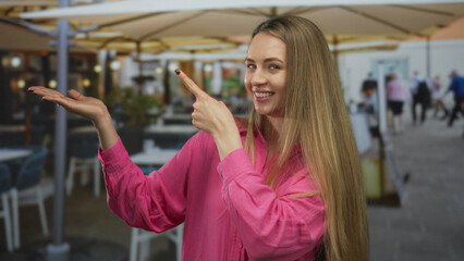 Fototapeta premium Woman smiling and pointing on a terrace at a restaurant with outdoor seating while wearing a pink shirt, showcasing a lively dining scene and casual ambiance.