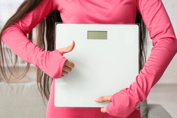 Beautiful young sporty woman with scales showing thumb-up gesture at home, closeup. Weight loss...