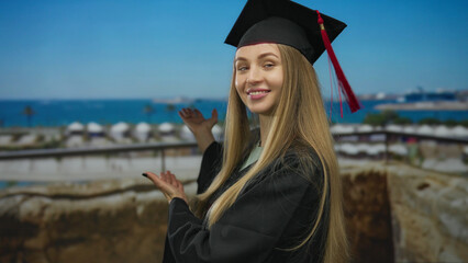 Woman in graduation cap and gown smiling at the camera with a scenic port and boat in the...