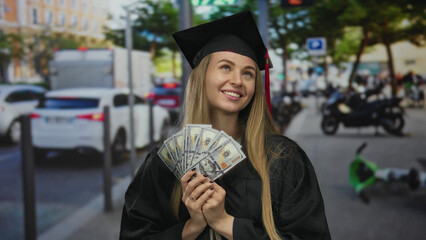 Woman smiling holding us dollars in graduation gown on city street, celebrating success, with...