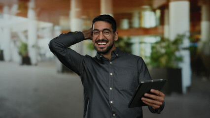 Young man with glasses smiles while looking at tablet in an urban setting, showcasing a modern, casual, and professional vibe in an outdoor environment.
