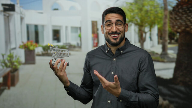 Man smiling outdoors in urban street holding toy shopping cart while wearing glasses and casual shirt.