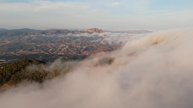 Drone video of Spanish mountains with dense clouds flowing over forested slopes, revealing peaks under clear blue sky.