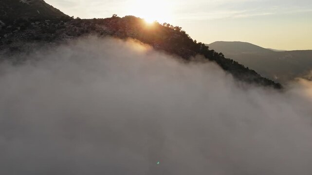 Drone video of sunset casting golden light over a forested Spanish mountain, with thick clouds and mist covering the slopes.