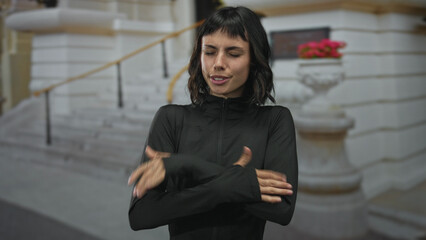 Young hispanic sporty woman in black jacket crosses arms over bare hands on building steps in cool outdoor weather; discomfort.