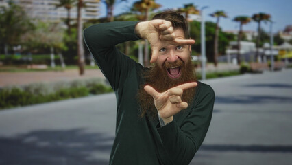 Man with long beard frames a scene with fingers while smiling on a city street in bright daylight;...