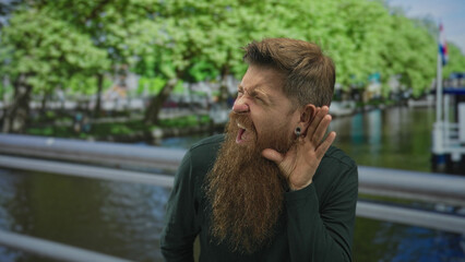 Man with red beard squinting and holding hand to ear on street railing by canal; curiosity...