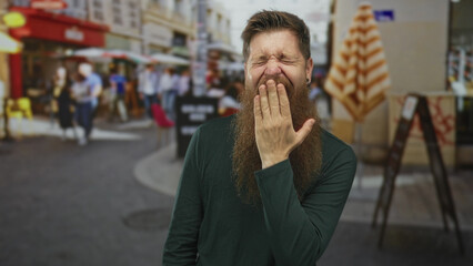 Bearded man in green sweater covers mouth with hand and squints eyes on a crowded city street;...