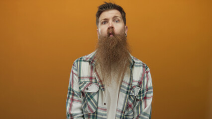 Man with long beard wearing checkered shirt making a pursed lip gesture in an orange studio; curiosity.