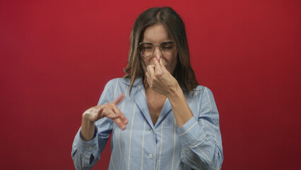 Young hispanic woman wearing glasses and striped shirt pinches nose in studio with red wall;...