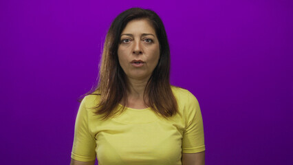 Hispanic middle aged woman covering mouth with hand in purple studio wearing yellow shirt and wide...