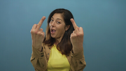 Middle aged hispanic woman showing middle finger gesture with both hands in blue studio setting;...