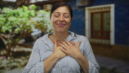 Middle aged hispanic woman holds hands to chest in green outdoor park beside blue building facade; gratitude calm.