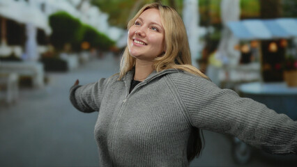 Woman smiling outdoors in city street wearing gray sweater, embodying joy and freedom, with relaxed demeanor in an urban setting as greenery and street vendors blur in the background.