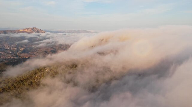 Drone video of Spanish mountains with dense clouds flowing over forested slopes, revealing peaks under clear blue sky.