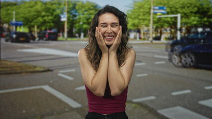 Young hispanic woman pressing cheeks with hands in a broad grin under sunlit trees on a busy street...