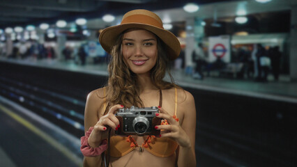Woman smiling in bikini holds camera at crowded indoor train station, wearing straw hat, representing young tourist at london railway platform scene.