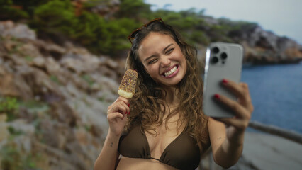 Woman enjoying ice cream while taking a selfie in swimwear at the beach with her smartphone,...