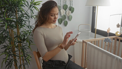 Pregnant woman using smartphone in stylish baby room with crib and plants, showcasing modern motherhood and preparation.