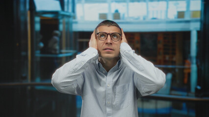 Obraz premium Man with glasses covering ears in hotel lobby, expressing stress and confusion amid modern indoor surroundings.