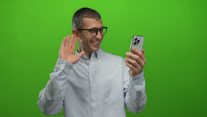 Man smiling at smartphone with green background engaging in video call showing cheerful expression and gesturing with hand while wearing glasses and casual shirt in isolated setting.