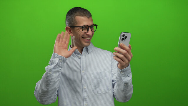 Man smiling at smartphone with green background engaging in video call showing cheerful expression and gesturing with hand while wearing glasses and casual shirt in isolated setting.