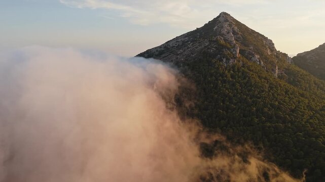 Drone video over Spanish mountain peak covered with forest, with golden sunset light illuminating drifting clouds in the foreground.