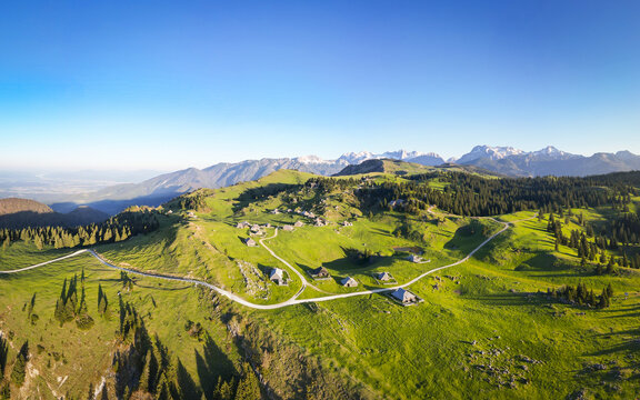 Aerial view of a lush green highland dotted with rustic wooden huts beneath a clear blue sky, contrasted by distant snow-capped mountain peaks, Stahovica, Kamnik, Slovenia.