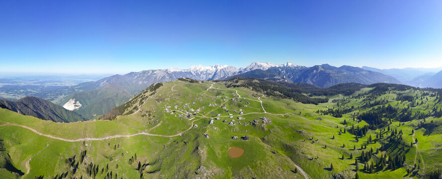 Aerial view of lush green pastures dotted with traditional shepherd's huts, set against a backdrop of snow-capped mountains, Stahovica, Kamnik, Slovenia.
