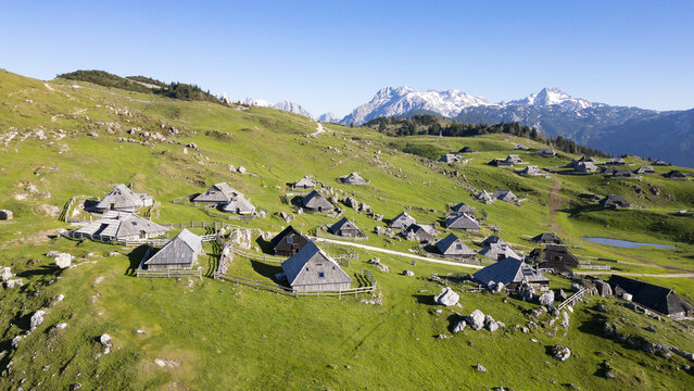 Aerial view of traditional wooden huts scattered across the vibrant green hillsides, framed by the majestic, snow-capped Alps, Stahovica, Kamnik, Slovenia.