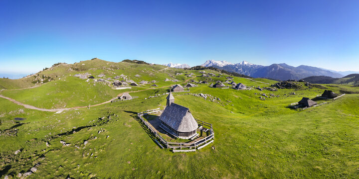 Aerial view of a wooden church with a tall spire stands serenely amidst verdant meadows, traditional huts dotting the landscape under a clear sky, Stahovica, Kamnik, Slovenia.