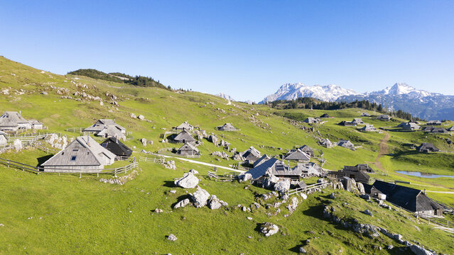 Aerial view of wooden huts scattered across vibrant green hillsides, contrasting with the distant snow-capped peaks, Velika Planina, Kamnik, Slovenia.
