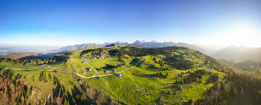 Aerial view of sun-drenched Velika Planina unfolds, with vivid green meadows dotted with wooden huts, and distant snow-capped mountains, Stahovica, Kamnik, Slovenia.
