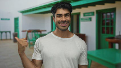 Young hispanic man pointing thumb behind him at a white house entrance on a sunny day; warm welcome.
