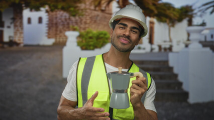 Young hispanic engineer in hardhat and neon vest holds moka pot above palm and smiles near...
