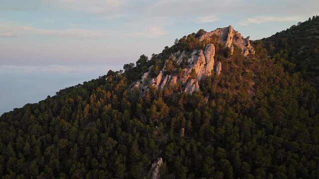 Aerial drone video of a forest-covered mountain peak in Spain at sunrise, with warm sunlight casting golden tones on rocky slopes.