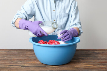 Woman in rubber gloves adding laundry detergent to clothes in plastic basin on wooden table against...