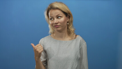 Young blonde woman pointing thumb over shoulder in blue studio setting with subtle smile; calm...
