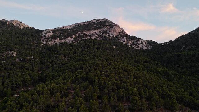 Aerial drone video of a forest-covered mountain peak in Spain at sunrise, with warm sunlight casting golden tones on rocky slopes.