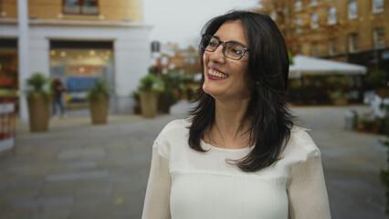 Woman wearing glasses smiling on city street with visible planters and outdoor cafe tables by...