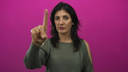 Middle age hispanic woman in olive sweater points index finger to camera in pink studio, wearing...