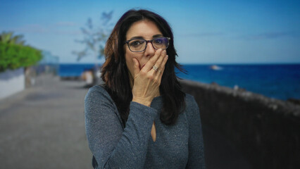 Middle aged woman with glasses making finger to lips gesture on a sunlit seaside promenade street;...