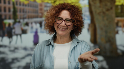 Woman with curly hair smiling outdoors in a city street, reaching out her hand with a cheerful expression against a blurred urban background.