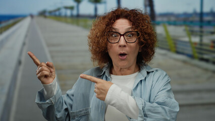 Middle-aged hispanic woman with curly brunette hair and glasses excitedly gesturing on a seaside...