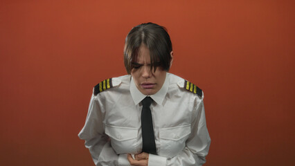 Woman pilot in uniform clutches stomach in discomfort against a red background, demonstrating a pained expression.